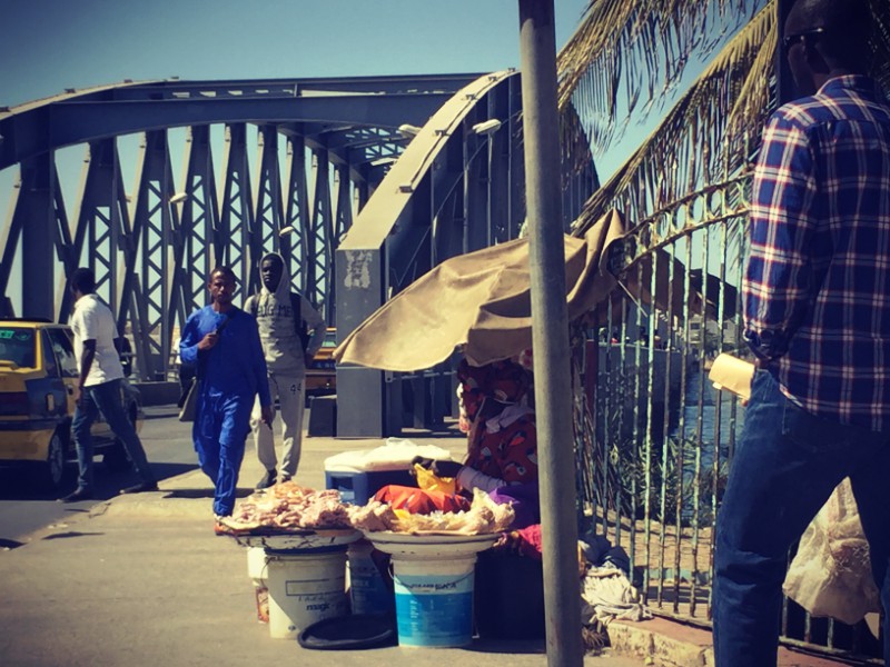 Des personnes marchent au bord du Pont Faidherbe, une marchande de fruits sous un parasol de fortune #Off2Africa 18 Nouakchott Saint-Louis © Gilles Denizot 2016