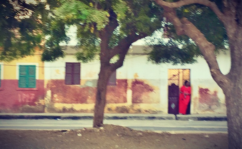 Une femme vêtue de rouge adossée contre sa porte, des arbres torsadés au 1er plan #Off2Africa 19 Saint-Louis Sénégal © Gilles Denizot 2016
