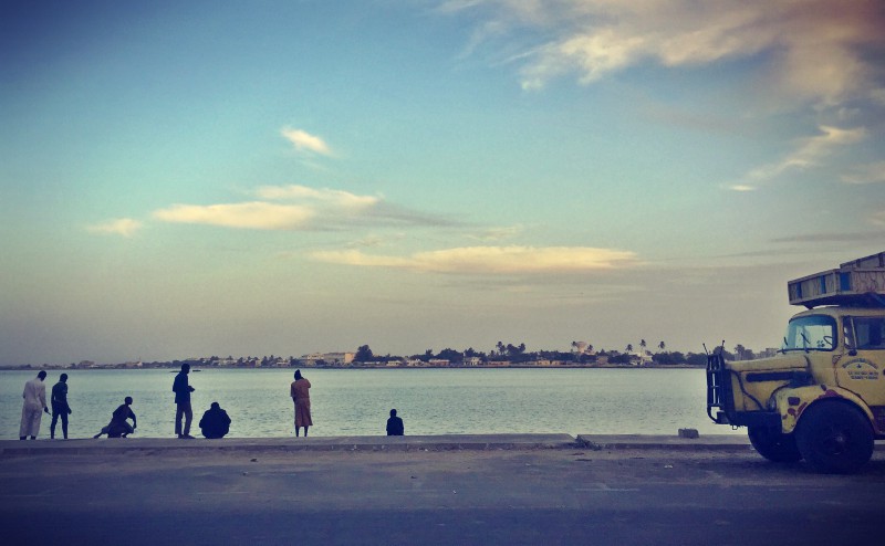 Vue sur le fleuve Sénégal. À droite, l'avant d'un camion jaune. À gauche, diverses silhouettes de personnes le long du fleuve #Off2Africa 22 Saint-Louis Sénégal © Gilles Denizot 2016