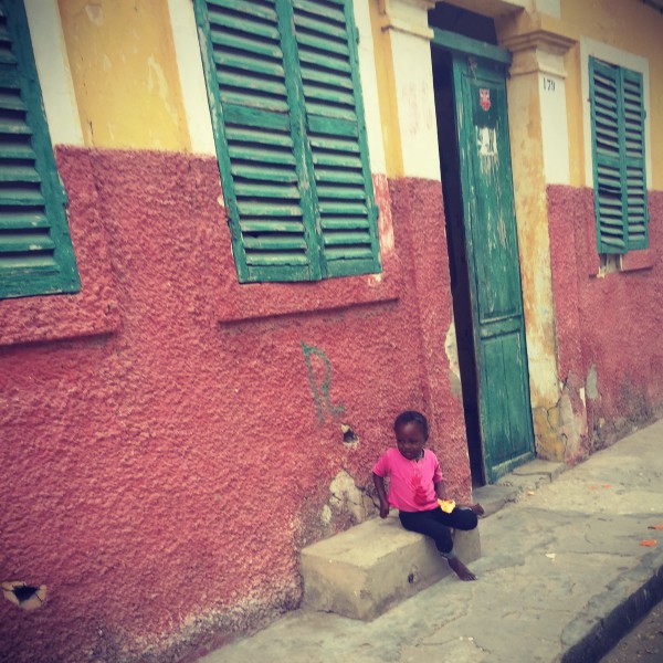 Une petite fille au tshirt rose assise devant une façade rose et jaune #Off2Africa 21 Saint-Louis Sénégal © Gilles Denizot 2016