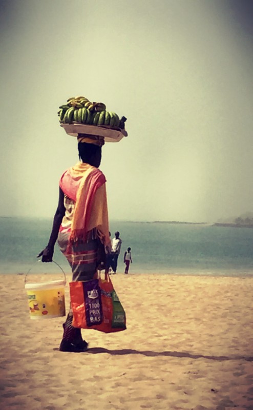 Une femme marche sur la plage. Elle charrie seau et sac, et porte un plateau jonché de bananes sur sa tête #Off2Africa 32 Mbour Sénégal © Gilles Denizot 2016