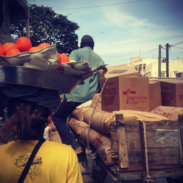 Embouteillage à Dakar, une femme avec dreads et t-shirt jaune porte un plat contenant des oranges sur sa tête, elle attend derrière une charrette sur laquelle un homme attend #Off2Africa 44 Dakar Ziguinchor Sénégal © Gilles Denizot 2017