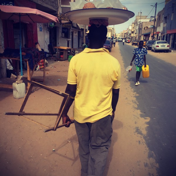 Un homme t-shirt jaune marche dans la rue ensablée et porte un plateau sur la tête #Off2Africa 44 Dakar Ziguinchor Sénégal © Gilles Denizot 2017