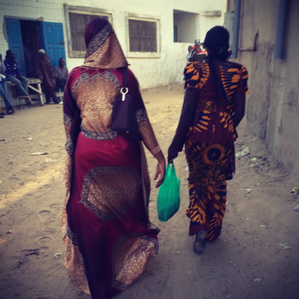 Deux femmes aux habits traditionnels colorés marchent dans une rue ensablée de Yoff. Des hommes assis sur la gauche #Off2Africa 39 Dakar Sénégal © Gilles Denizot 2017