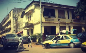 Coin de rue aux maisons jaunies, une voiture jaune et bleue et deux hommes marchant #Off2Africa 64 Conakry Guinée © Gilles Denizot 2017