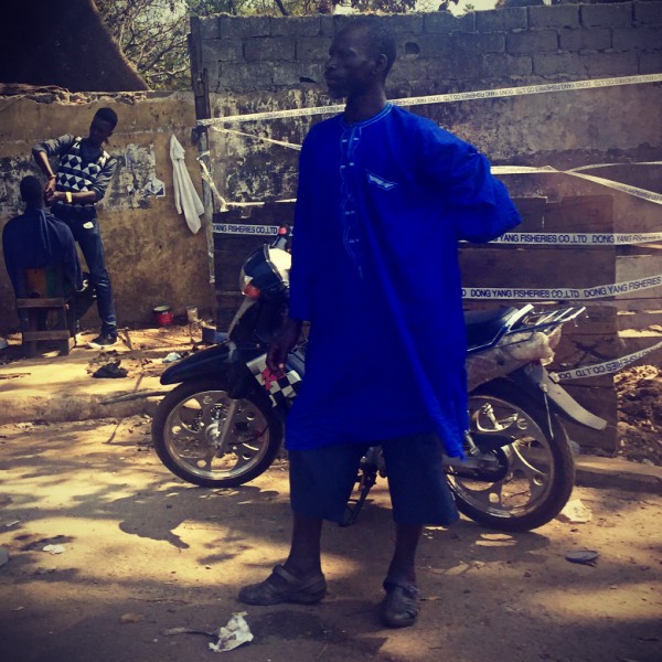Un homme vêtu de bleu attend debout, derrière lui s'affaire un barbier #Off2Africa 54 Conakry Guinée © Gilles Denizot 2017