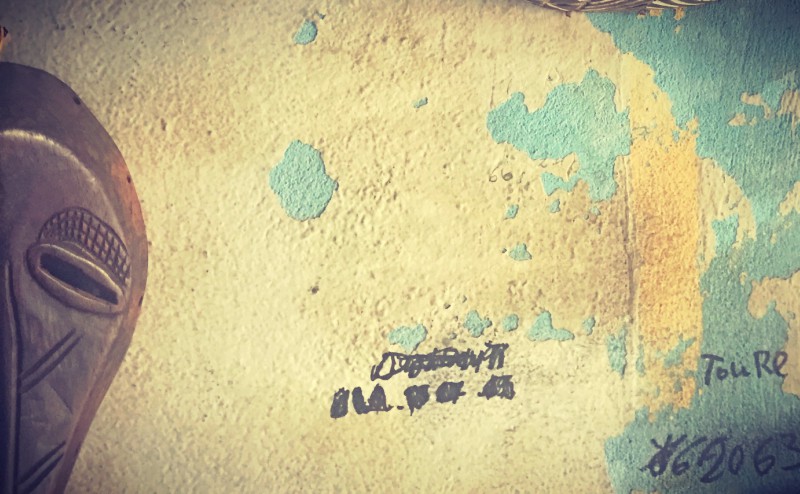 Une partie d'un masque africain en bois contre un mur décrépi bleu et blanc, quelques inscriptions au feutre noir #Off2Africa 77 Conakry Guinée © Gilles Denizot 2017