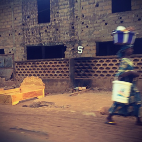 Dans une rue ensablée de Bamako, une femme portant des seaux sur la tête et au bras marche vivement. Un immeuble en construction et devant un cadre de lit en bois #Off2Africa 85 Bamako Mali © Gilles Denizot 2017