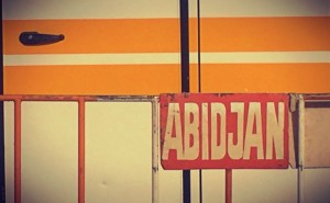 Sur une barrière rouge et blanche, un écriteau rouge indique ABIDJAN. Derrière, le détail d'un autobus orange #Off2Africa 91 Abidjan Côte d'Ivoire © Gilles Denizot 2017