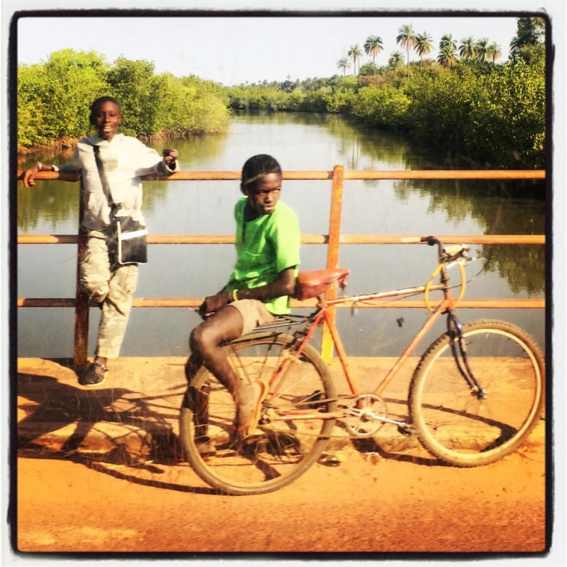 Un garçon chemise verte sur l'arrière d'un vélo, un autre sweat-shirt blanc accoudé sur la rambarde d'un pont #Off2África 49 Ziguinchor Bissau © Gilles Denizot 2017