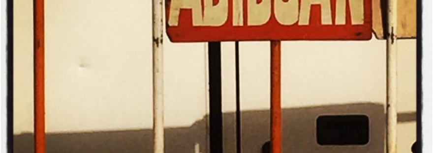 Sur une barrière rouge et blanche, un panneau indique ABIDJAN. Derrière, le détail d'un autobus #Off2Africa 91 Abidjan Côte d'Ivoire