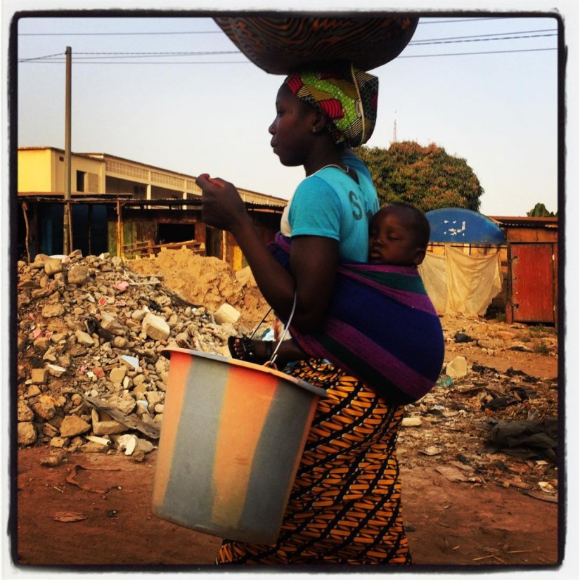 Dans une rue de Bamako, un femme porte un ballot sur la tête, un enfant dans le dos et un seau au bras gauche #Off2Africa 85 Bamako Mali © Gilles Denizot 2017