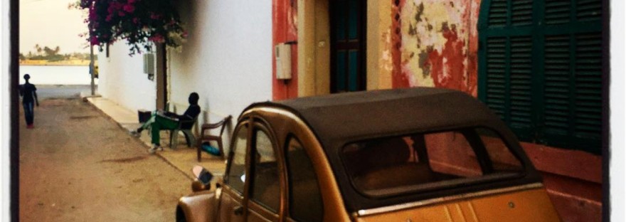 Une Citroën 2CV couleur bronze parquée dans une ruelle de Saint-Louis, contre une façade ocre décrépie. Plus loin un homme assis sur un banc, sous une immense bougainvillée et encore plus loin un homme debout. Vue sur le fleuve au loin. #Off2Africa 22 Saint-Louis Sénégal © Gilles Denizot 2016
