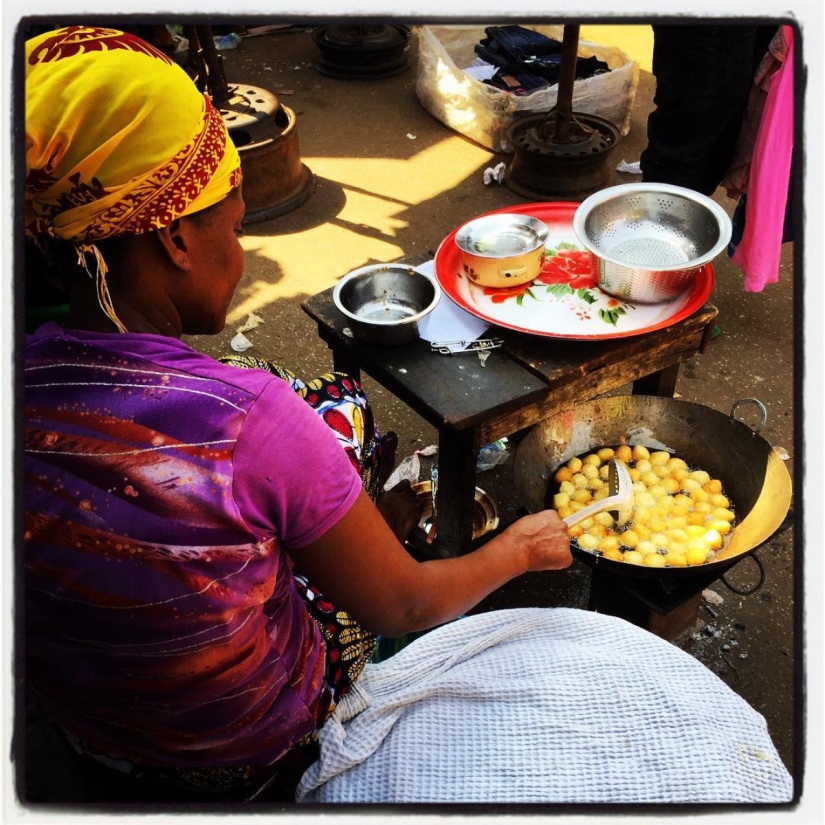 Assise devant un réchaud, une femme vêtue d'un t-shirt violet, la tête couverte d'un foulard jaune. De la main droite, elle remue des beignets ronds dans l'huile du réchaud #Off2Africa 73 Conakry Guinée