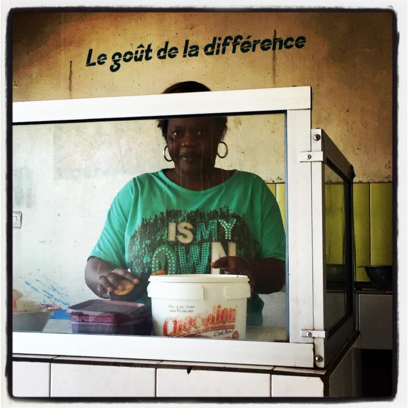 Une femme derrière une vitre. Sur le mur est écrit : Le goût de la différence #Off2Africa 37 Dakar Sénégal © Gilles Denizot 2017