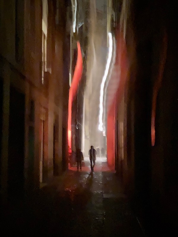 Dans une rue du Raval, deux silhouettes marchent entre des lueurs rouges et blanches #HolaBCN Elémentaire Vicenç © Gilles Denizot 2022