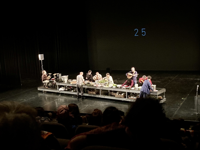 Une grande table sur une scène, des personnes cuisinent autour © Gilles Deniozt 2023