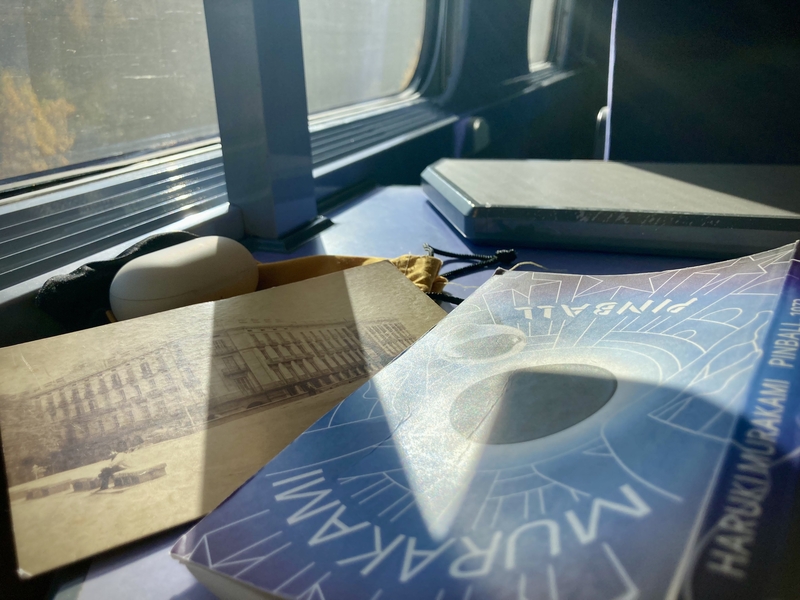 Sur une tablette de train, une carte postale d'un vieux bâtiment de Barcelone et la couverture de Pinball de Murakami #Off2Europe Resfeber Stress © Gilles Denizot 2023