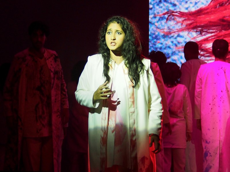 Photo de la production #ItsComplicated, Chennai, Inde. Eurydice vêtue de blanc, derrière elle se tiennent des chanteurs, l'un de face, les autres de dos sur fond rougeâtre ©GD14 #HolaBCN